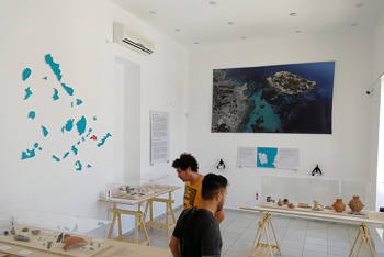 Two men observe the exhibit at the Museum of Koufonisi, which includes a collection of pieces of broken Cycladic figurines